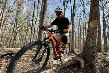 A cyclist in a black t-shirt and knee pads rides an orange mountain bike on a rugged trail surrounded by trees. The scene captures the rider navigating a rocky path, with bare branches visible in the background under a clear blue sky. Locust Shade Park mountain bike trail.