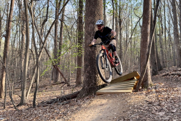 A mountain biker in mid-air jumps off a wooden ramp in a forested area, surrounded by tall trees and fallen leaves. The biker is wearing a helmet and protective gear, showcasing an action-oriented moment in outdoor sports. Locust Shade Park mountain bike trail.