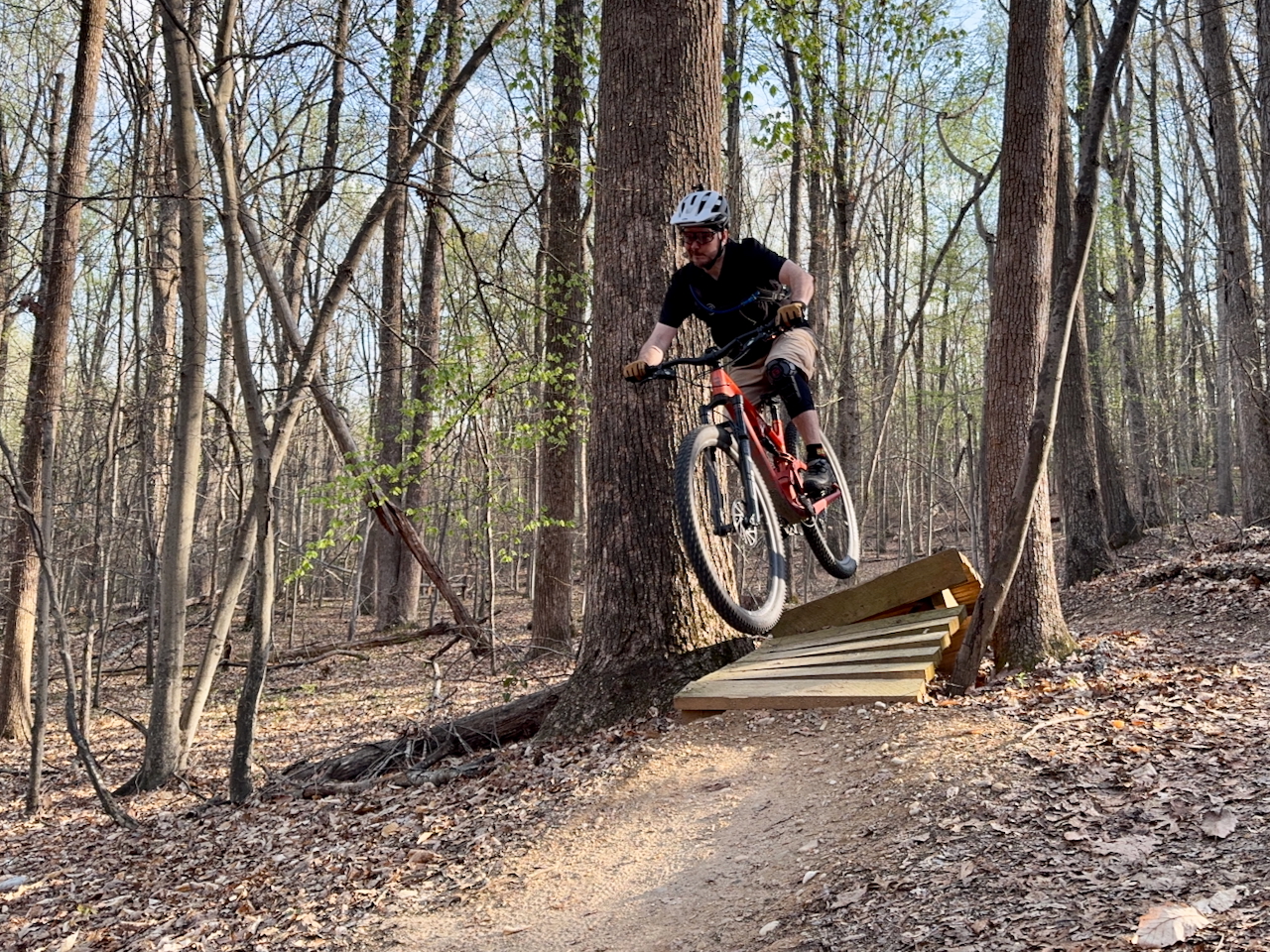 A mountain biker in mid-air jumps off a wooden ramp in a forested area, surrounded by tall trees and fallen leaves. The biker is wearing a helmet and protective gear, showcasing an action-oriented moment in outdoor sports. Locust Shade Park mountain bike trail.