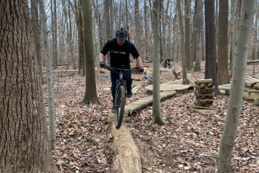 A mountain biker skillfully rides along a narrow log bridge in a wooded area, surrounded by bare trees and fallen leaves. The cyclist wears a helmet and a black shirt, focused on maintaining balance while navigating the challenging terrain. In the background, another biker can be seen approaching a wooden structure on the trail. Locust Shade Park mountain bike trail.