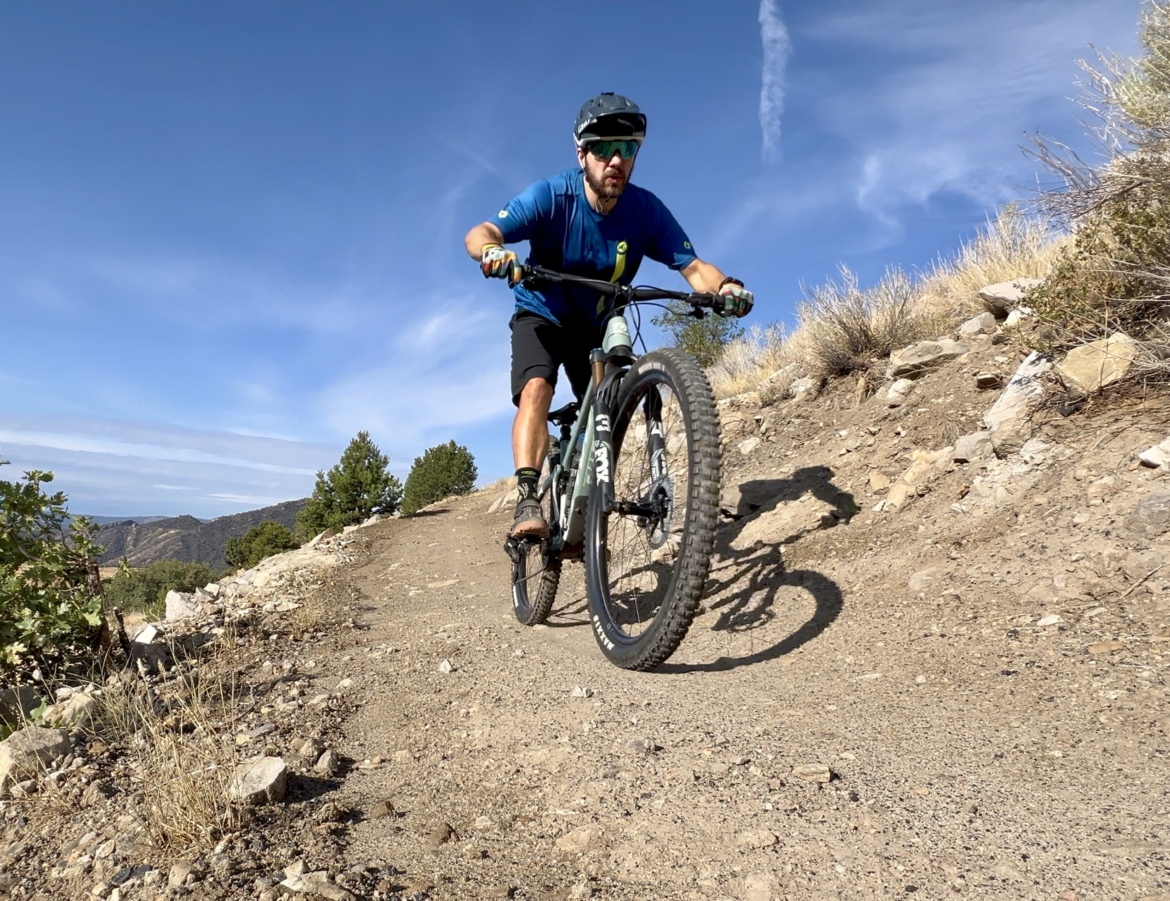 A cyclist riding a mountain bike on a rocky trail, surrounded by shrubs and trees under a blue sky with scattered clouds. The cyclist is focused and wearing a helmet and sunglasses. The trail is dusty, showing signs of wear from biking activity.