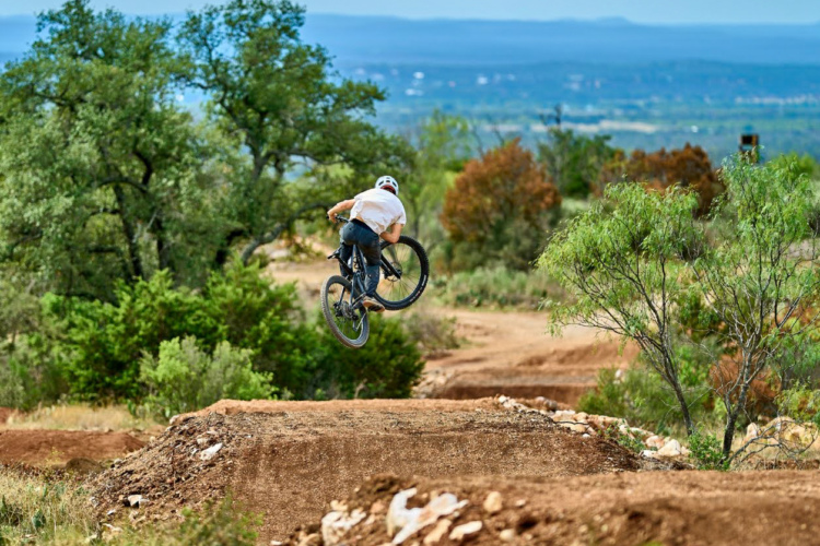 A mountain biker performing a jump over a dirt ramp, surrounded by greenery and a distant landscape. The rider is wearing a helmet and casual clothing, with trees and brush evident in the background under a clear blue sky.