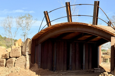 A curved metal bridge with a wooden deck, set against a clear blue sky. The bridge is partially constructed and framed with support beams, surrounded by rocky terrain and sparse trees in the background. Rock Yard mountain bike trail.