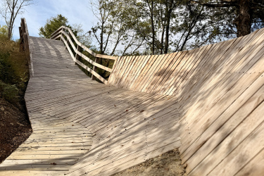 A wooden ramp with a curved design, surrounded by trees. The ramp features a sloped surface with a railing along one side, leading into a natural area under a bright sky. Rock Yard mountain bike trail.