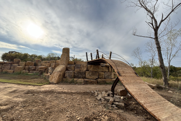 A wooden bridge arching over a rocky landscape, connecting two elevated areas. The background features a large stone structure and a barren tree against a cloudy sky, with the sun partially visible. The ground is dirt and grass, with scattered rocks. Rock Yard mountain bike trail.