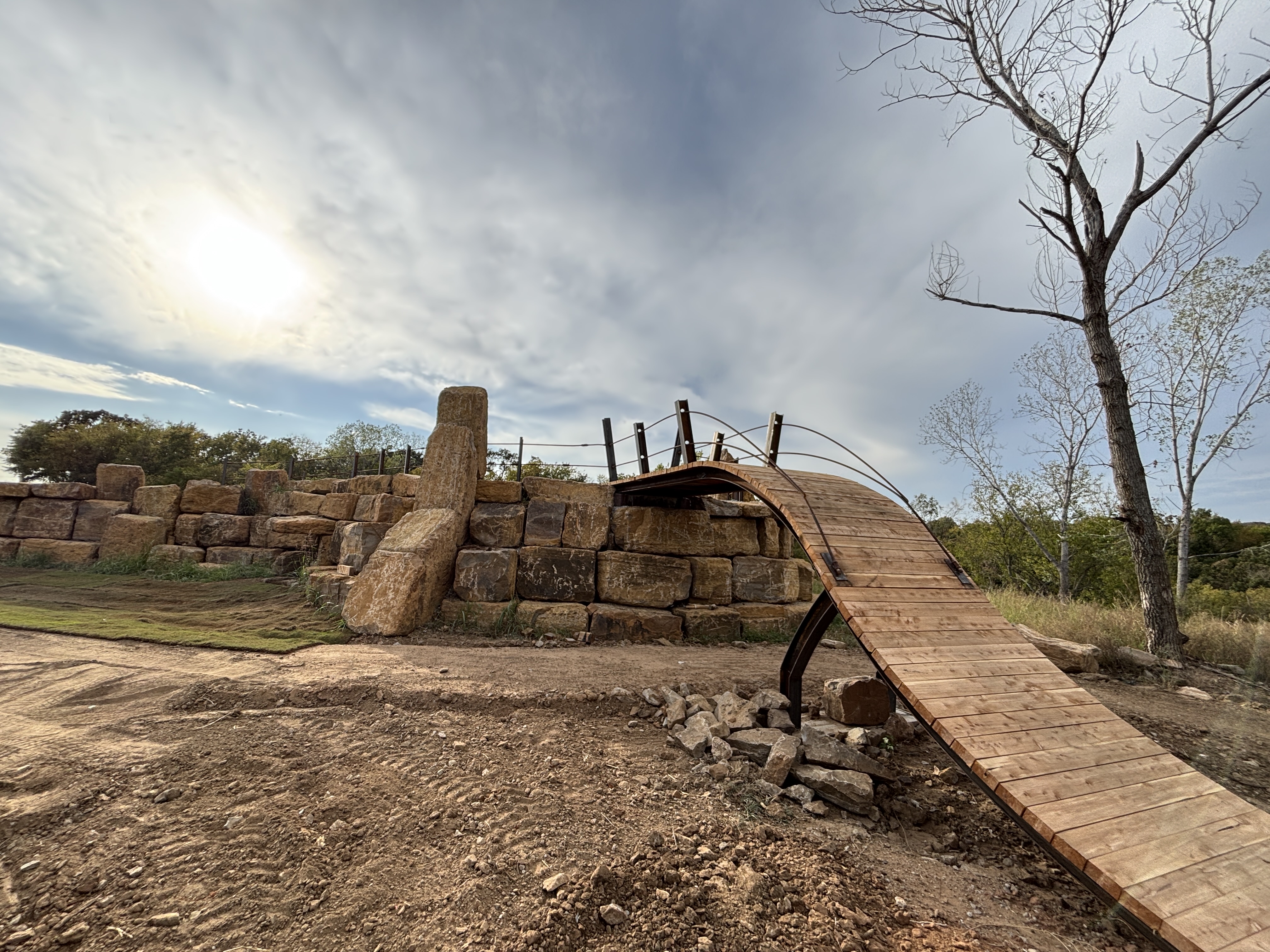 A wooden bridge arching over a rocky landscape, connecting two elevated areas. The background features a large stone structure and a barren tree against a cloudy sky, with the sun partially visible. The ground is dirt and grass, with scattered rocks. Rock Yard mountain bike trail.