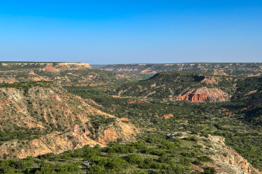 A panoramic view of a rugged canyon landscape featuring layered red and beige rock formations, lush greenery, and vast blue skies. The scene captures the natural beauty of the terrain with rolling hills and distant cliffs under a clear horizon. Palo Duro Canyon mountain bike trail.