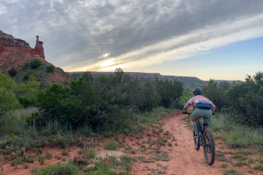 A mountain biker riding along a dirt trail in a canyon landscape during sunset. The path is surrounded by green shrubs and rocky formations, with a prominent red rock structure visible in the background under a cloudy sky. Palo Duro Canyon mountain bike trail.