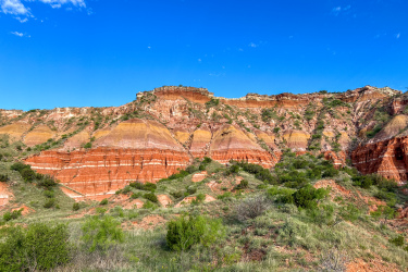 A vibrant landscape featuring layered red and orange rock formations against a clear blue sky. The foreground showcases green shrubs and grass, providing a contrast to the colorful cliffs in the background. Palo Duro Canyon mountain bike trail.