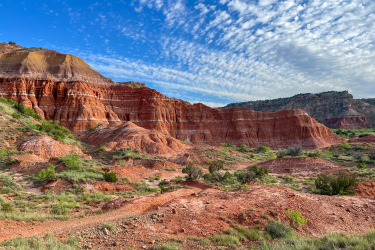 A scenic view of rugged red rock formations and rolling hills under a blue sky with wispy clouds, featuring patches of green vegetation in the foreground and on the slopes. The landscape showcases the natural beauty of a desert environment. Palo Duro Canyon mountain bike trail.