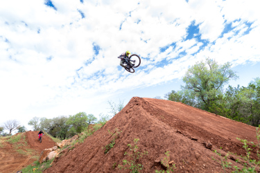 A mountain biker performing a jump over a dirt ramp against a backdrop of blue skies with scattered clouds. Another biker is visible in the background, riding on a different dirt track surrounded by greenery. Reveille Peak Ranch mountain bike trail.