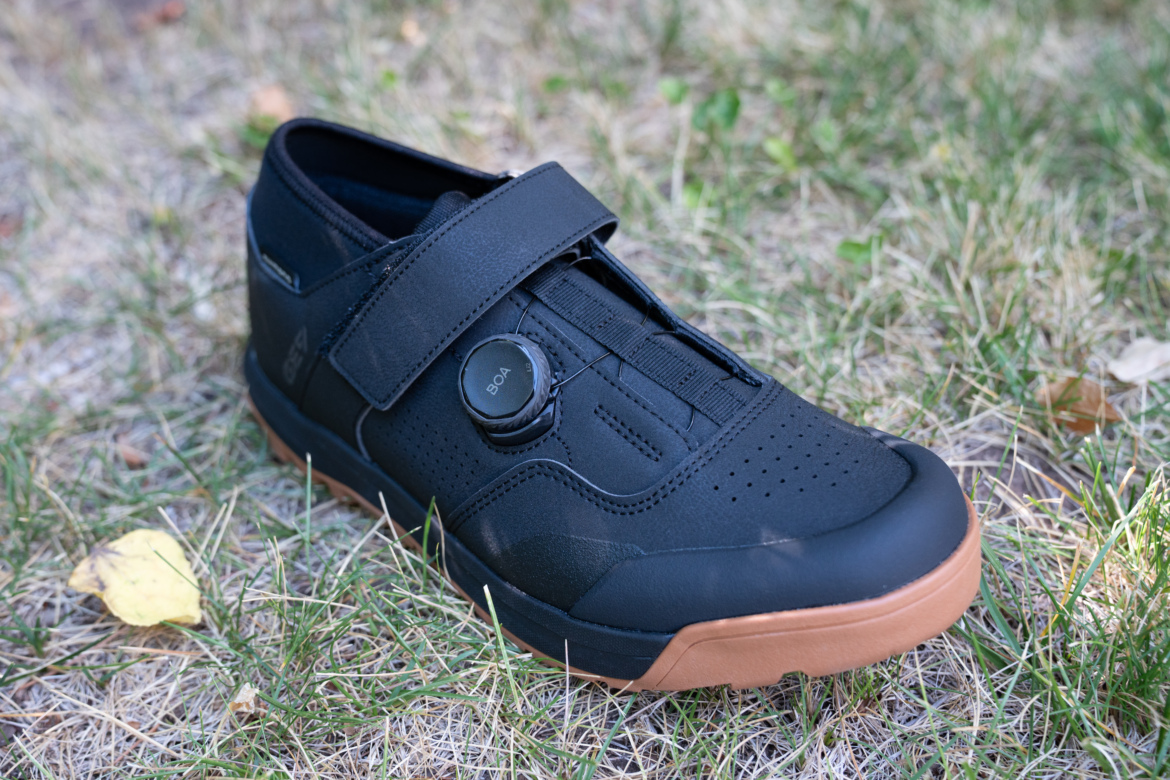 A black athletic shoe with a textured surface, featuring a velcro strap and a circular dial closure system, resting on dry grass alongside a small yellow leaf.