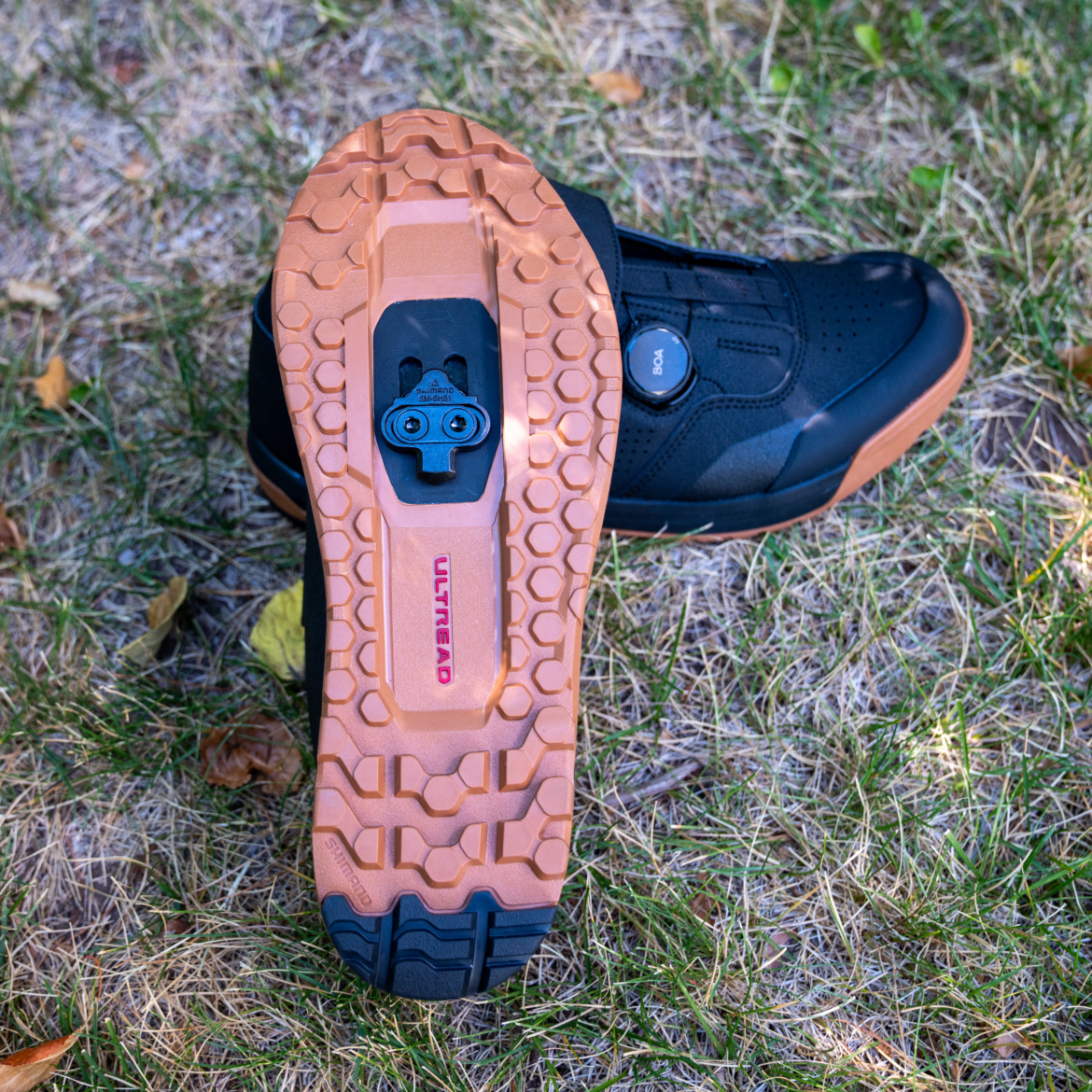 A close-up view of the sole of a black cycling shoe featuring a multi-textured rubber outsole with a light brown tread pattern. The shoe has a clip mechanism visible in the center, along with a BOA dial for tightening. The background consists of dry grass and scattered leaves, providing a natural outdoor setting.