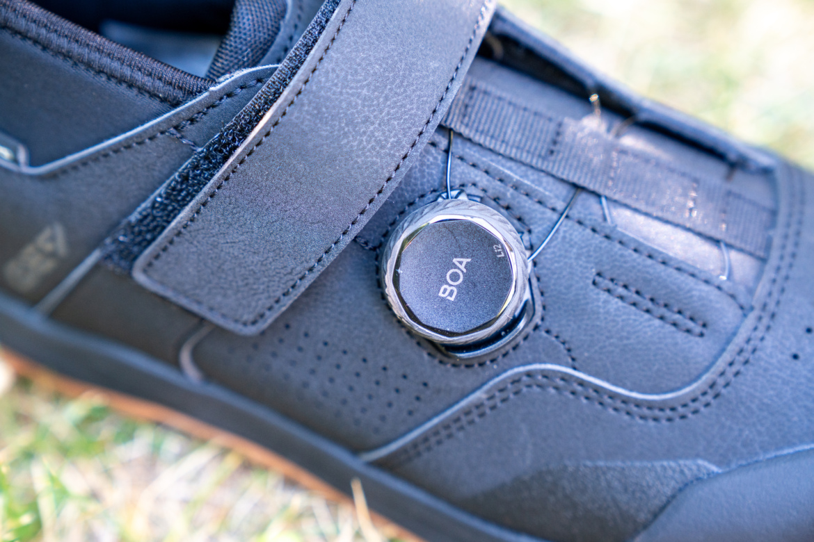 Close-up of a black cycling shoe featuring a BOA lacing system. The shoe has a sleek design with perforations for ventilation and a Velcro strap for added security. The focus is on the BOA dial, which is used for quick adjustments to the fit. The background is blurred grass, emphasizing the shoe's details.