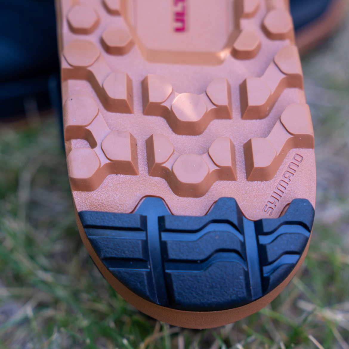 Close-up view of the sole of a shoe, featuring a tread pattern with hexagonal and rounded shapes, primarily in shades of brown and black. The sole has a logo engraved on the side, indicating the brand is Shimano. In the background, a blurred grassy surface is visible.