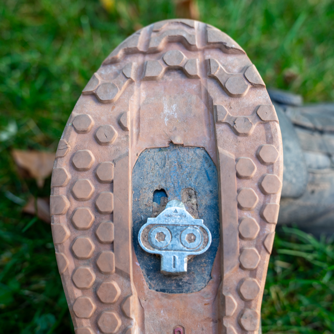 Close-up view of a cycling shoe's sole, featuring a clip for attaching to a bike pedal. The sole is textured with a hexagonal pattern and shows signs of wear, with a brown and gray color scheme against a backdrop of green grass.