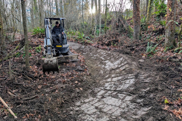 A person operating a small excavator to reshape a muddy trail in a forested area, surrounded by trees and fallen leaves. The scene captures the equipment in action, with a focus on the newly modified path.