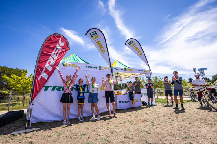 A group of young cyclists celebrating on a podium after a race, displaying medals and trophies. They are surrounded by banners from Trek and NICA, with spectators and volunteers in the background. The sky is clear and blue with a few clouds, hinting at a bright, sunny day.