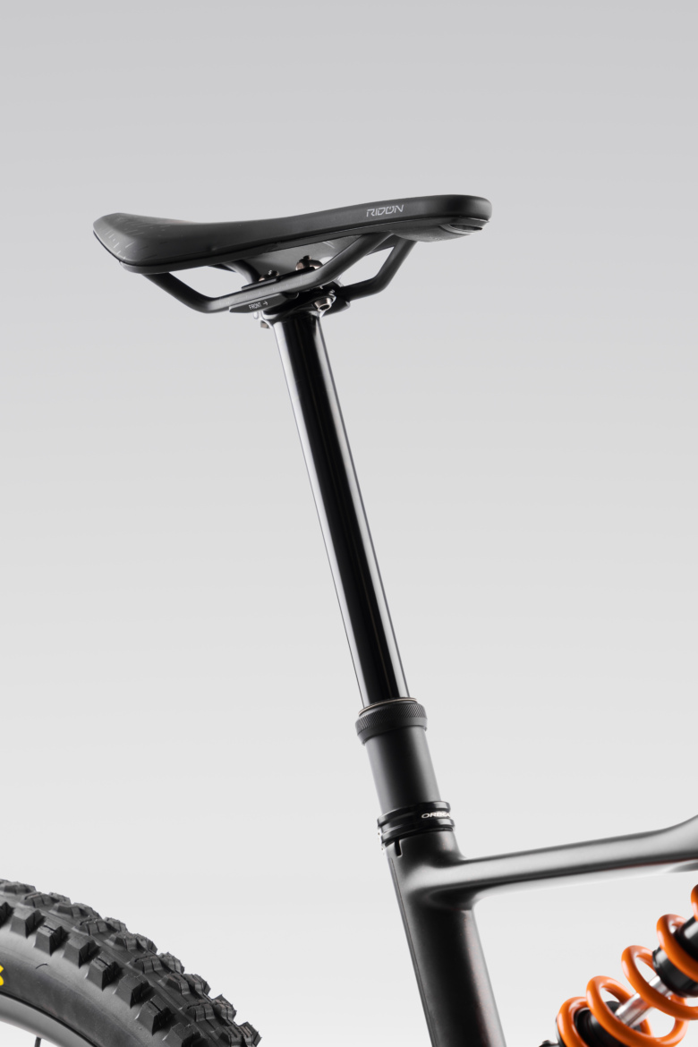 Close-up image of a mountain bike seat and post, showcasing a sleek black design and an orange suspension spring. The seat has a modern shape, with the post extending upward, set against a neutral gray background.