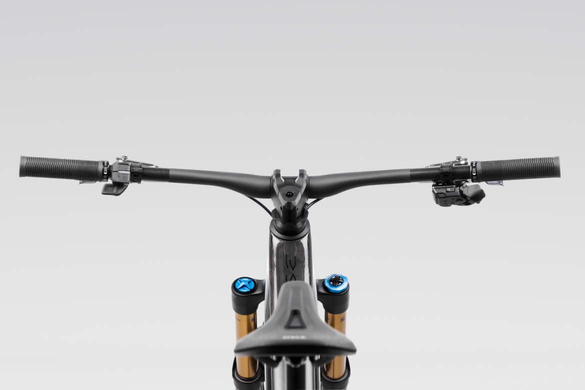 A close-up view of a mountain bike's handlebars, featuring textured grips, brake levers, and gear shifters. The handlebars connect to a sleek, black frame, with gold suspension struts visible below. The background is a soft gradient, emphasizing the bike's components.