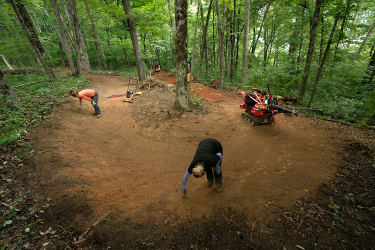 Two people are working on a dirt path in a wooded area. One person is bending down to clear debris from the ground, while the other is using a small machine for landscaping. Surrounding them are tall trees and lush greenery, with tools and equipment visible in the scene. Aetna Mountain mountain bike trail.