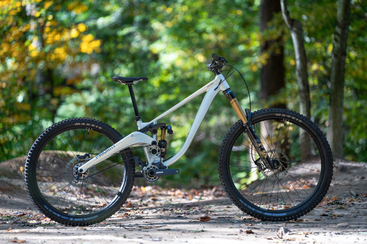 A modern mountain bike with a white frame and gold suspension fork, placed on a dirt trail surrounded by greenery and autumn leaves. The bike features large, knobby tires and a single chainring setup, showcasing its design for rugged terrain.