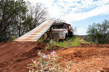 An old, rusty white pickup truck parked on a dirt path, with a wooden ramp leading up to it. Surrounding the truck are green shrubs and trees, under a partly cloudy sky. In the foreground, a few colorful wildflowers are visible on the dirt. Reveille Peak Ranch mountain bike trail.