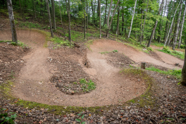 A dirt bike trail winding through a wooded area, featuring multiple banked turns and natural landscaping. The ground is covered with loose dirt and pebbles, surrounded by green vegetation and trees. Mt. Telemark mountain bike trail.