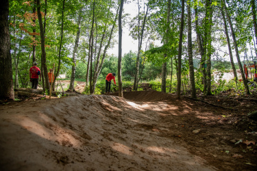 Two individuals work on a dirt bike trail in a forested area. One person, wearing an orange shirt, stands near a tree while the other, dressed in a red shirt, is seen digging in the dirt on a section of the trail. The surroundings are lush with greenery and sunlight filtering through the trees, highlighting the natural setting. Mt. Telemark mountain bike trail.