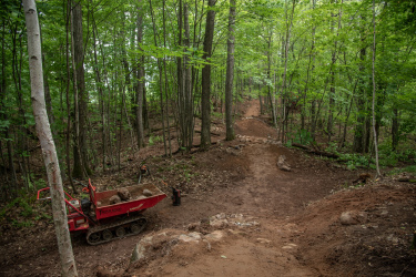 A forested area with green trees and a dirt path where construction or trail maintenance is taking place. In the foreground, a small red tracked dumper is loaded with rocks, while a trail winds up the slope in the background. The scene illustrates a blend of nature and outdoor construction work. Mt. Telemark mountain bike trail.