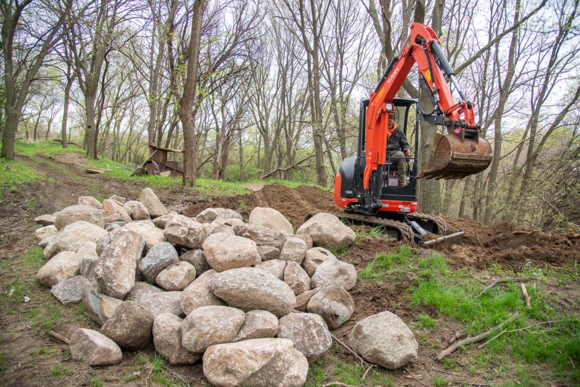 A small excavator is prominently featured in a forested area, digging into the earth near a large pile of variously sized rocks. In the background, a path winds through the trees, and an old structure is visible. The scene captures a mix of construction and natural elements, highlighting the ongoing landscaping or excavation work.