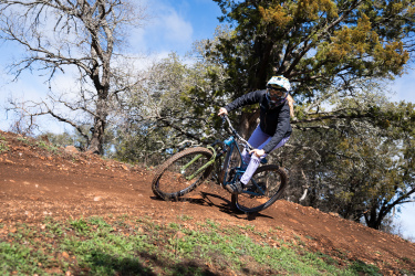 A mountain biker riding along a dirt trail, leaning into a sharp turn. The rider is wearing a helmet and cycling gear, with trees in the background and a blue sky overhead. Reveille Peak Ranch mountain bike trail.