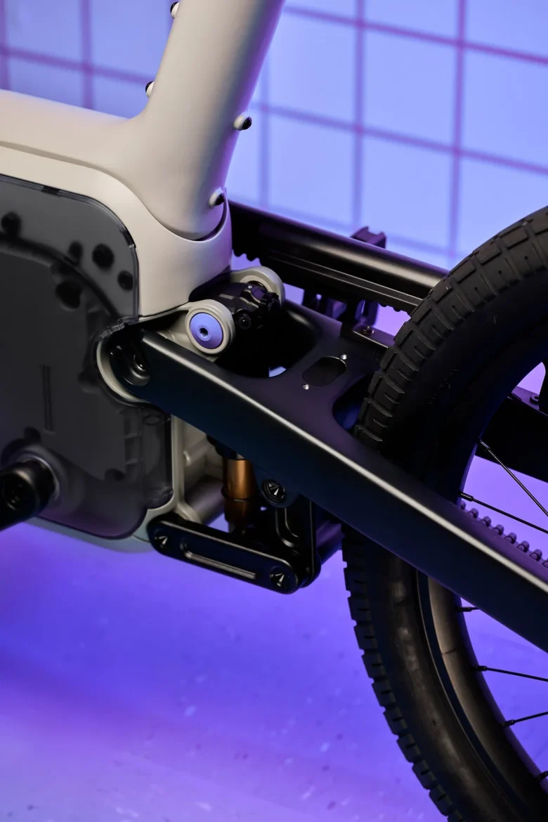 Close-up of a modern bicycle's drivetrain and frame, showcasing the intricate components and design. The image features a blend of metallic and matte textures with a dark background and subtle purple lighting.