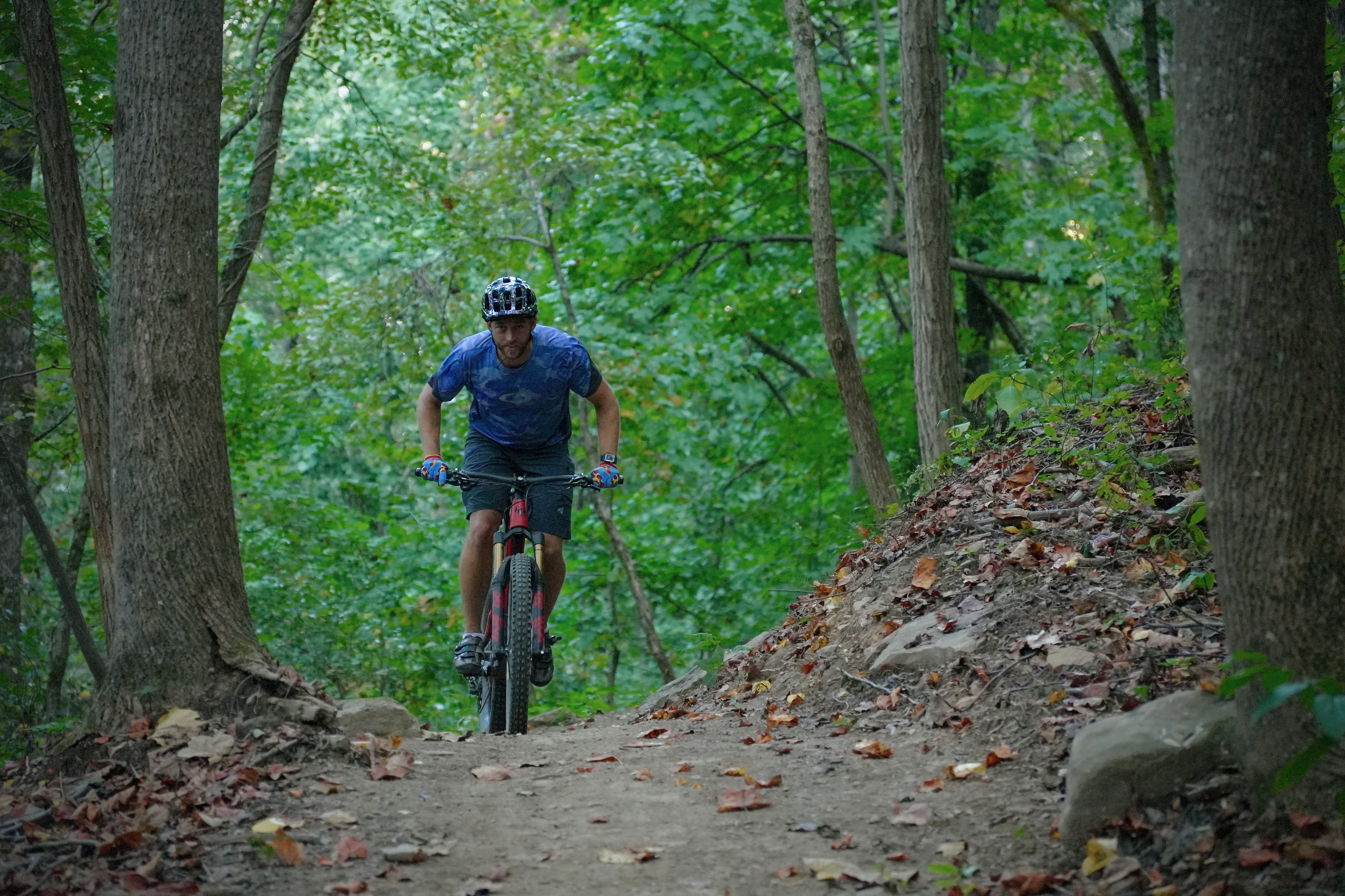 A mountain biker navigating a dirt trail surrounded by lush green trees, with autumn leaves scattered on the ground. The cyclist is wearing a helmet and is positioned in an active riding posture. Aetna Mountain mountain bike trail.
