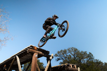 A person in a black helmet and riding gear is performing a jump on a turquoise mountain bike, soaring above a wooden obstacle in a clear blue sky. Trees are visible in the background, indicating an outdoor biking trail or park. Reveille Peak Ranch mountain bike trail.
