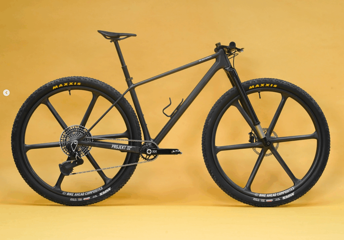 A sleek black mountain bike with unique large, five-spoke wheels and a lightweight frame, set against a bright yellow background. The bike features Maxxis tires, a chain drive system, and a minimalist design, emphasizing its performance-oriented features.