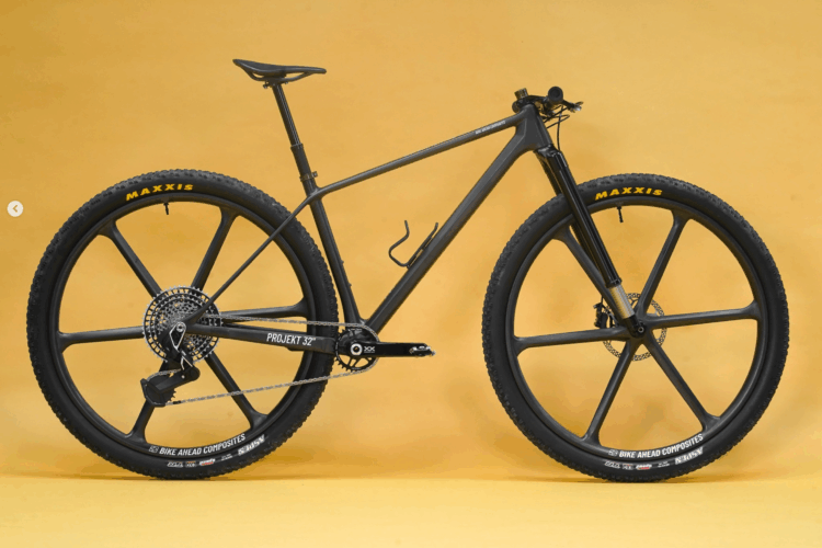 A sleek black mountain bike with unique large, five-spoke wheels and a lightweight frame, set against a bright yellow background. The bike features Maxxis tires, a chain drive system, and a minimalist design, emphasizing its performance-oriented features.