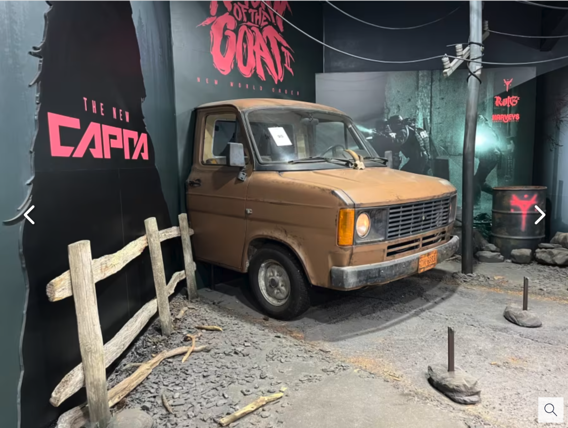Alt text: A vintage brown van positioned against a dark wall featuring bold red text that reads "THE NEW CAPTA." The scene includes a wooden fence, a barrel with graffiti, and an industrial aesthetic with scattered rocks and a dimly lit atmosphere.