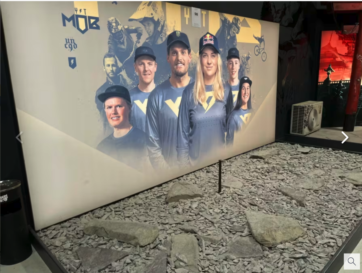 A large display features a group of six athletes, showcasing their team spirit with matching blue jerseys and caps. The background displays action shots of various extreme sports. The foreground includes a rocky terrain, resembling a training or competition setup, with scattered stones and gravel.