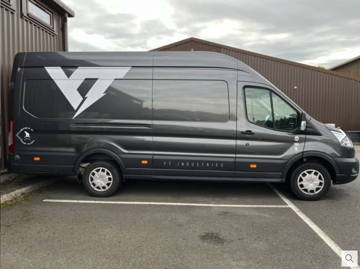 A gray delivery van with the logo "YT" prominently displayed on the side, parked in a lot. The van has a sleek design and is positioned next to a building with overhead windows. The sky is overcast in the background.