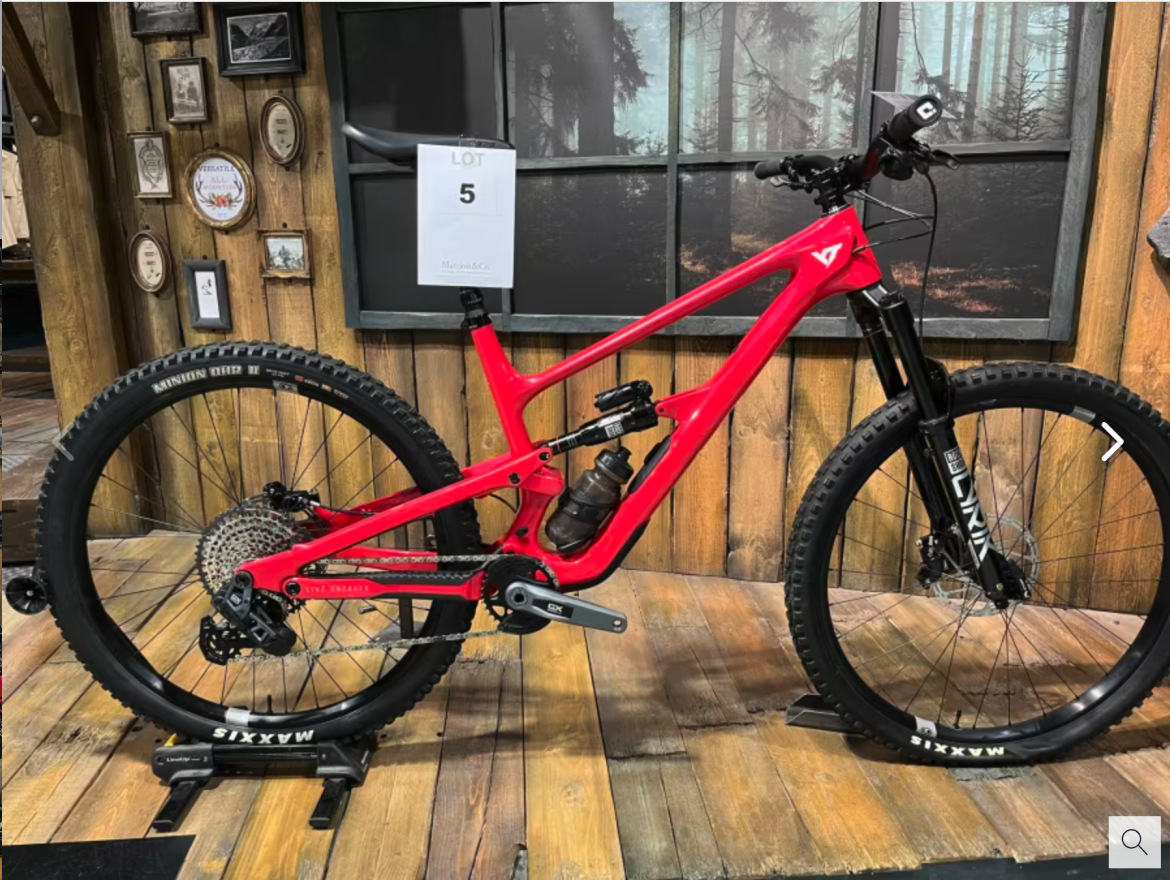 A vibrant red mountain bike displayed against a wooden backdrop, featuring a modern frame design, thick tires, and a water bottle mounted on the frame. The display includes a sign labeled "LOT 5."