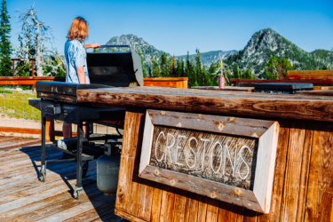 A person standing near an outdoor grill on a wooden deck with mountains and lush trees in the background. The grill is open, and the area features a wooden sign that reads "CRESTONS." A beverage can is visible on the counter. The scene is set in a clear, sunny environment. Anthony Lakes Mountain Resort mountain bike trail.