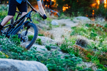 A young individual wearing a helmet and a short-sleeved shirt rides a mountain bike over rocky terrain, with a vibrant sunset and blurred trees in the background. Anthony Lakes Mountain Resort mountain bike trail.