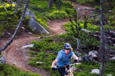 A mountain biker riding on a dirt trail through a lush green forest, with the sunlight filtering through the trees. The biker is in a dynamic position, leaning into the turn, and kicking up dust behind the bike. Anthony Lakes Mountain Resort mountain bike trail.