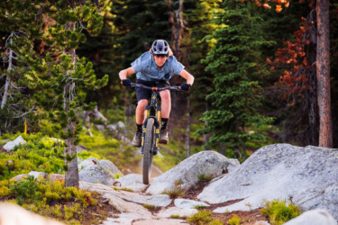 A mountain biker in a helmet and casual attire jumps over rocky terrain, surrounded by lush greenery and tall trees. The rider is focused, showcasing an adventurous spirit as they navigate the challenging trail. The image captures the thrill and excitement of mountain biking in a natural setting. Anthony Lakes Mountain Resort mountain bike trail.