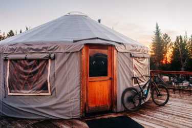 A cozy yurt with a wooden door and a bike parked beside it, set against a backdrop of trees and a vibrant sunset sky. The yurt features a grey exterior and a few windows, indicating a tranquil outdoor setting. Anthony Lakes Mountain Resort mountain bike trail.