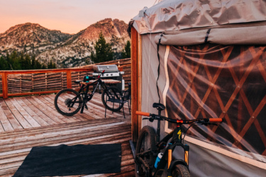 A wooden deck overlooking mountainous terrain at sunset, featuring a yurt with a clear wall, a bicycle parked nearby, and a grill set up for outdoor cooking. Anthony Lakes Mountain Resort mountain bike trail.