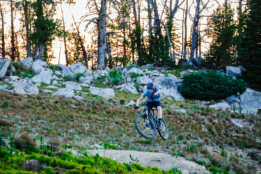 A person riding a mountain bike performs a jump over a rocky terrain, surrounded by trees and vegetation. The scene captures the dynamic movement and excitement of mountain biking in a natural outdoor setting during sunset. Anthony Lakes Mountain Resort mountain bike trail.
