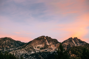 Sunset view of rugged mountain peaks against a colorful sky, with patches of trees in the foreground and hints of fading light in the background. Anthony Lakes Mountain Resort mountain bike trail.