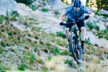 A person in a helmet and gloves is performing a jump on a mountain bike while riding down a rocky slope. The background features trees and rocky terrain, capturing the excitement of outdoor biking. The scene is set during sunset, with warm light illuminating the landscape. Anthony Lakes Mountain Resort mountain bike trail.